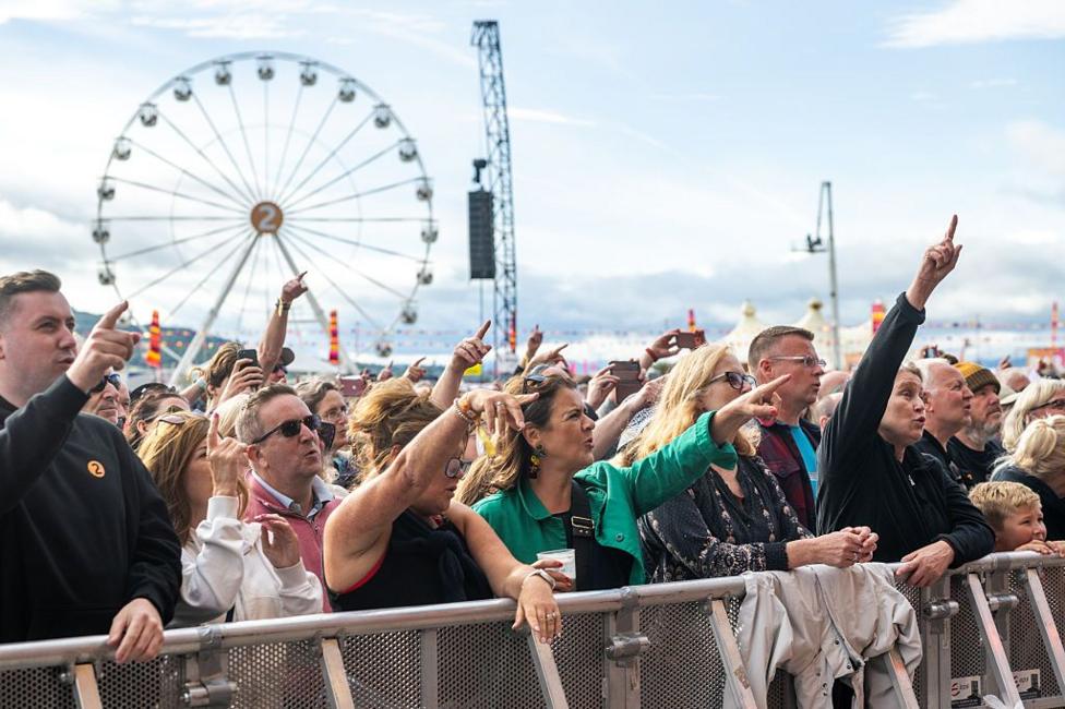 Crowd of people at a festival. A number of them are pointing towards the stage. There is a big wheel behind them.