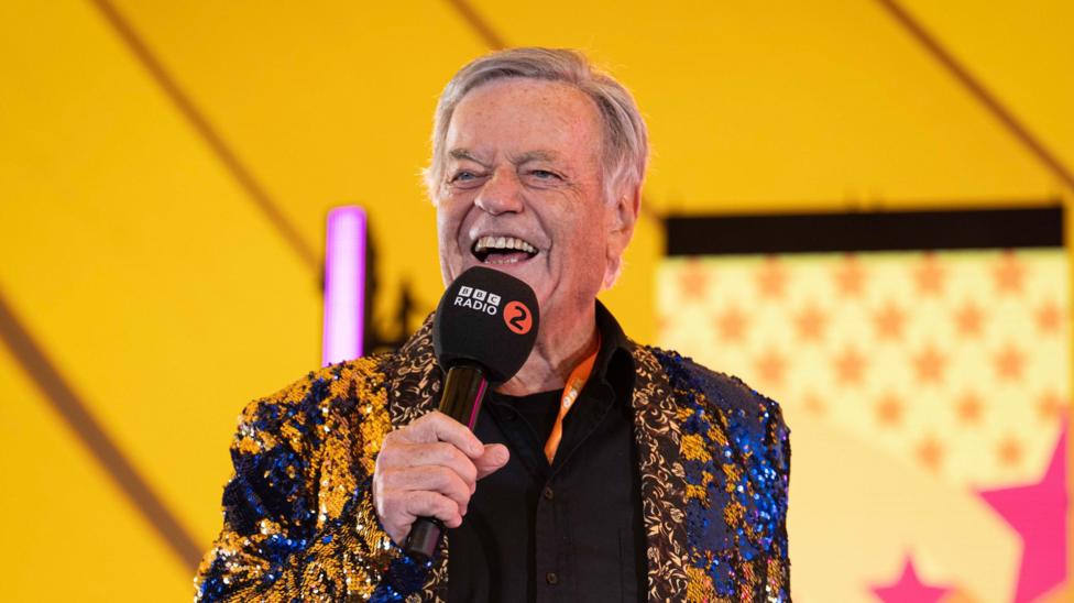Radio 2 DJ Tony Blackburn addresses the crowd on stage. He is smiling and wearing a gold and blue sparkly jacket