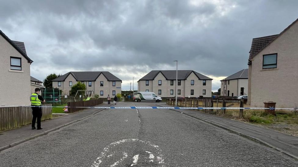 Blue and white police tape stretched across a road with a uniformed officer in a hi-vis jacket standing on the pavement. There are a number of houses in the picture as well as vehicles and children's play equipment in a garden