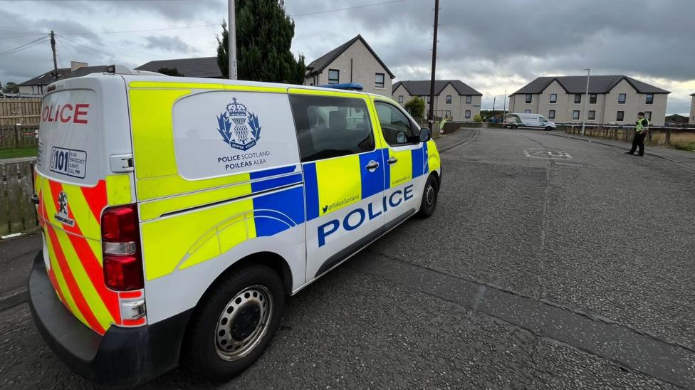 A police van parked in a street with an officer further on, standing next to police tape that is stretched across the road.