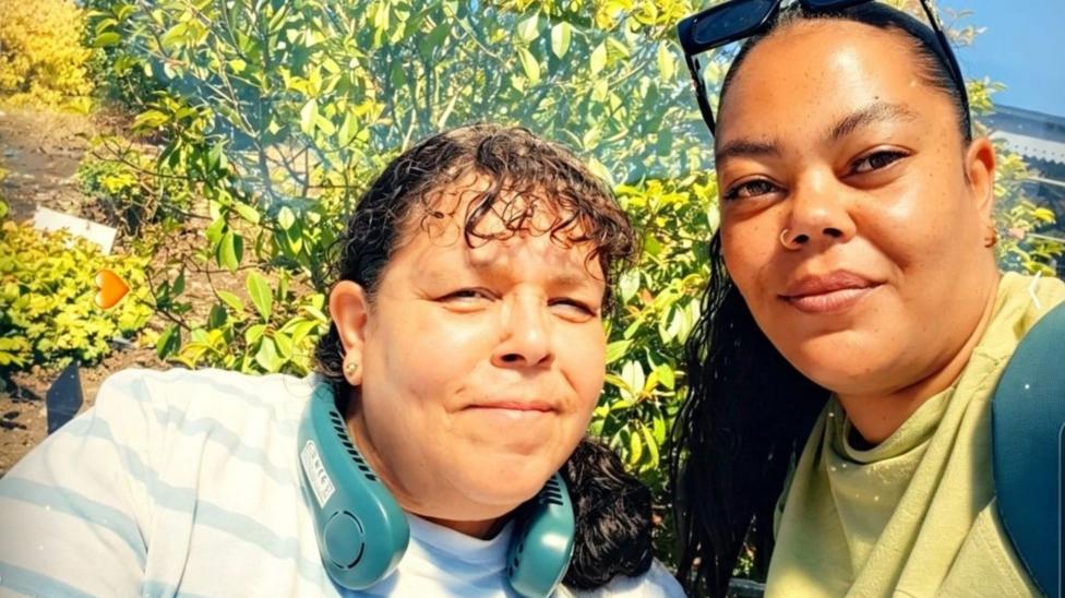 A selfie photograph of Natalie (right) and her mum (left). Natalie has sunglasses on top of her head and smiles at the camera. She has a nose ring and brown eyes. Her mother wears a blue and white striped top, a blue neck fan, and smiles at the camera. She has brown curly full fringe and a ponytail. 