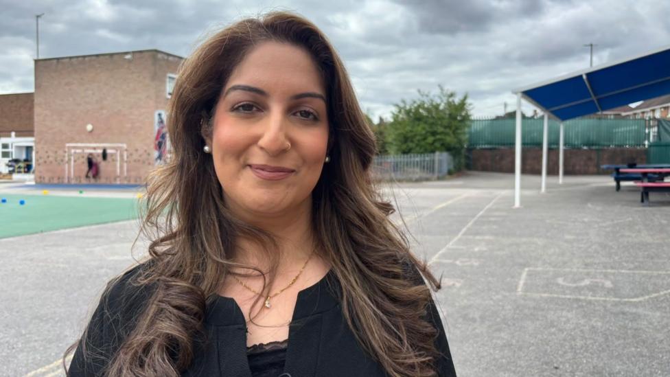 Farzana has long dark hair wearing a black top and necklace, standing in a school playground with a brick building, covered seating area, and cloudy sky in the background.
