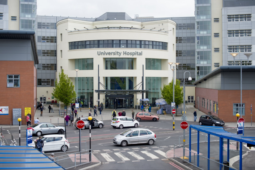 The exterior of University Hospital in Coventry. It has a semi circular entrance with windows and a wide door, with red brick buildings either side. Cars are parked outside the entrance