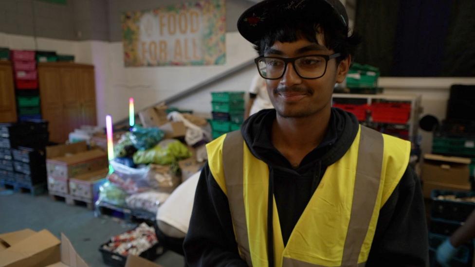 A young man in a high-visibility vest and cap stands in a warehouse with crates of food behind him.