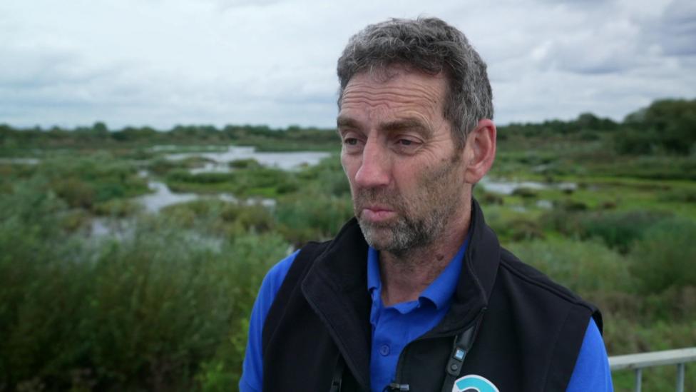 A man with grey hair is wearing a blue polo shirt and black gilet. He is standing in front of wetlands.