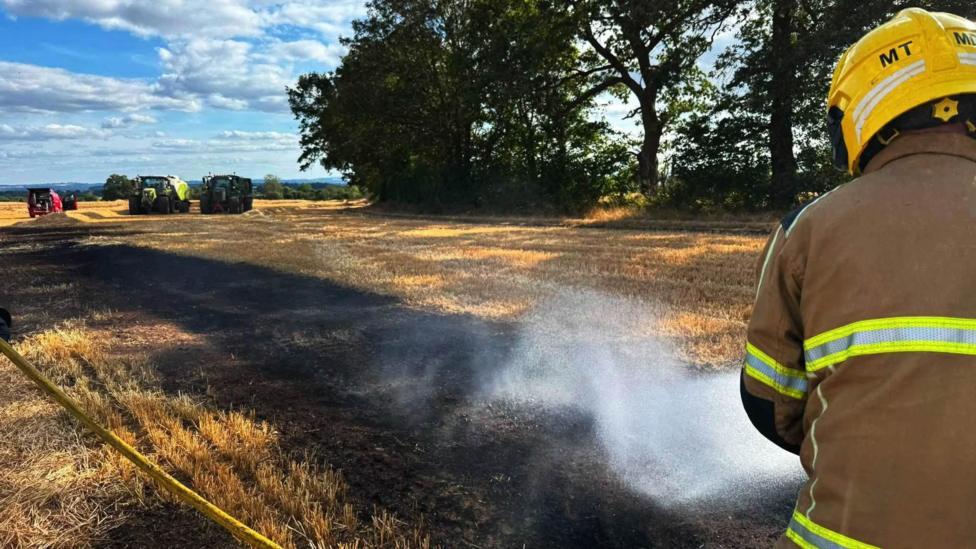 A firefighter with a yellow helmet and brown overalls is spraying a charred area of crops with water. the crops have been damaged by fire. in the background there are farm vehicles including a green tractor 