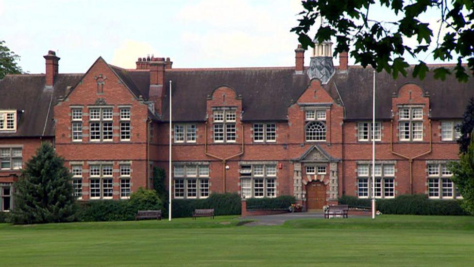 Exterior of a university which is a large, red-brick building with two storeys. In front of the university is a green lawn with two white flagpoles. 
