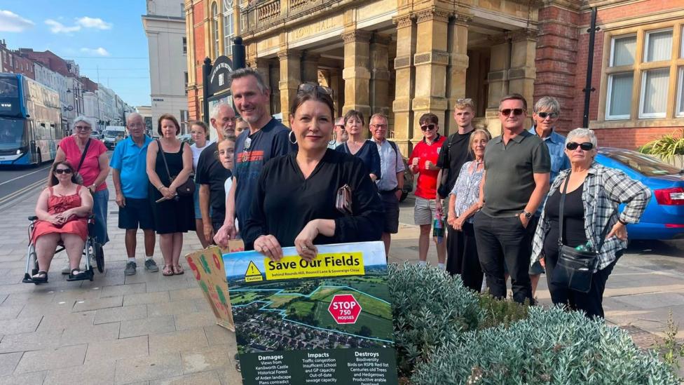 A group of people stood outside a town hall building, in a city centre street, holding a save our fields sign