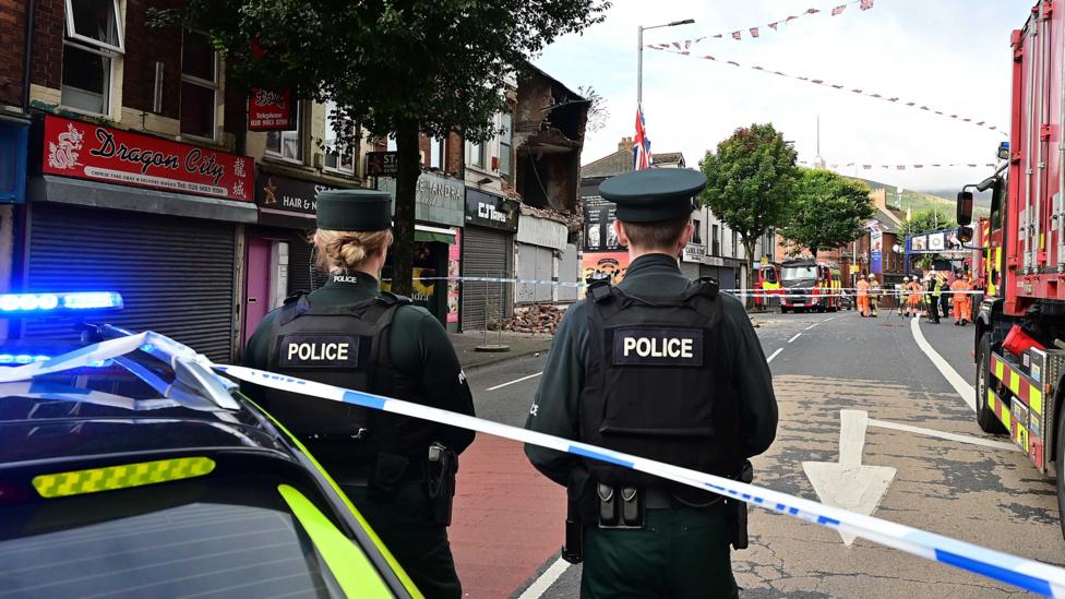 Two police officers standing in front of a partially collapsed building. The roof of a police car is visible. The road is cordoned off with blue and white police tape.
