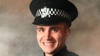 A headshot of a policeman, smiling for the camera while in uniform with his black police hat on.