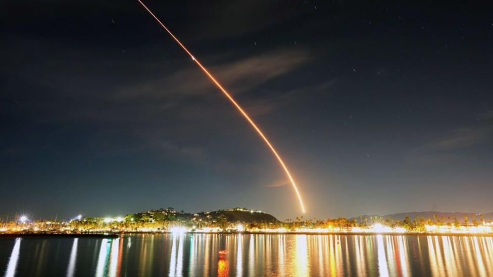 The photo shows a rocket being launched in the dark night sky. There is an orange trail that follows it. Lots of lights from buildings line the bottom of the photo.