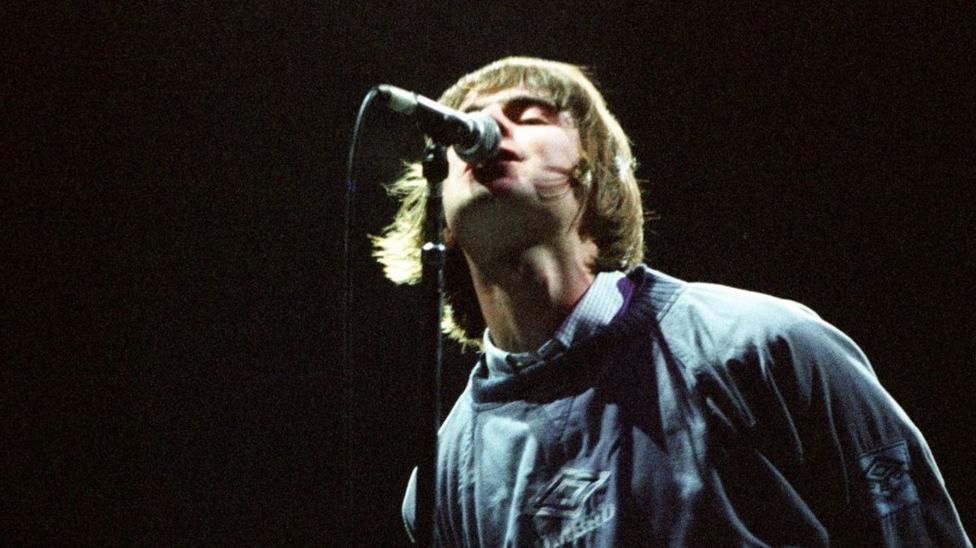 Liam Gallagher wearing an Umbro jumper while singing into a mic during one of Oasis's famous Maine Road gigs in 1996