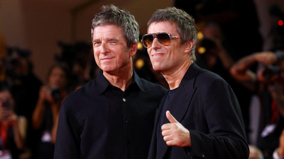 Noel and Liam smiling while on the red carpet at the Venice Film Festival ahead of the premiere of their comeback tour film, Don't Look Back in Anger. Liam is wearing shades and giving a thumbs up