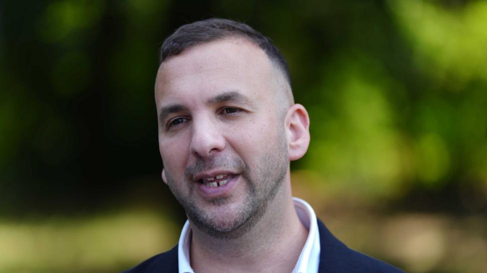 A man in a dark suit and white shirt with dark hair and stubble is pictured with some foliage out of focus in the background.