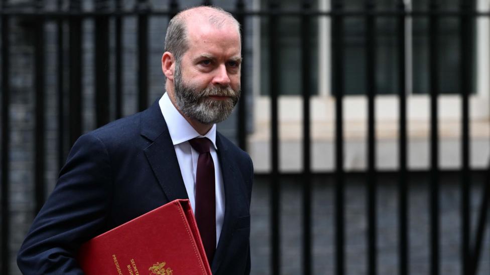 Business secretary Jonathan Reynolds carrying a red folder against the backdrop of some black, iron railings. He is wearing a dark suit and white shirt.