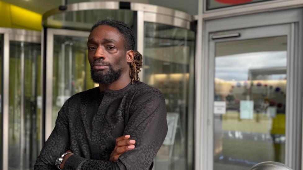 Efeotor with arms crossed, dressed all in black, with beard and tied back dreadlocks, looks straight at the camera with a serious expression.