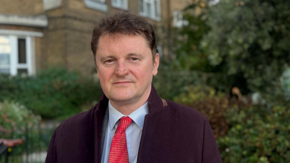 Davies pictured looking straight at the camera, wearing a light blue shirt, red tie and dark overcoat. He has short hair and is clean-shaven.