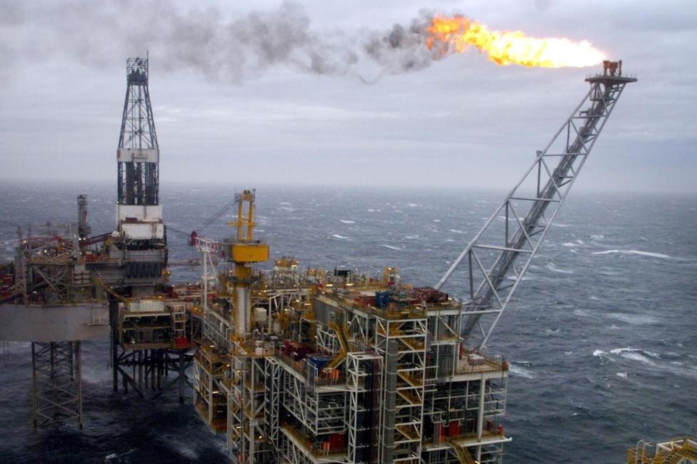 An offshore oil platform in rough seas with a bright flare on top of a diagonal stack. The platform is attached to a drilling rig.