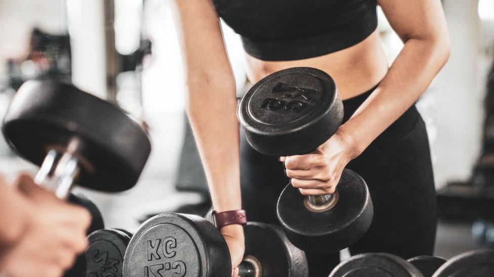 A woman in black leggings and a top lifts black dumbells in a gym