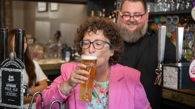 Wearing a pink blazer, the Welsh government's finance minister Elin Jones sips a freshly-poured pint behind the bar at the Radyr Tap. There is a man with a beard and glasses, with a black shirt stood smiling behind her.
