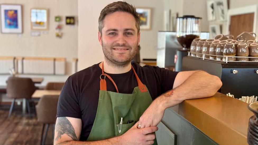 A man in a nave tshirt and green apron leans one arm on a high counter, with restaurant seating in the background