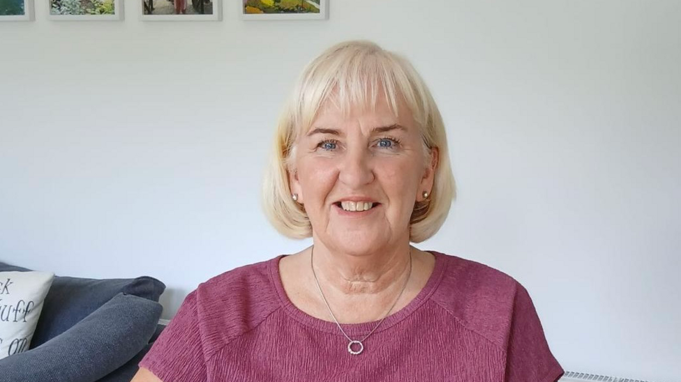 Amanda Pollock, a woman with grey/blonde hair, wearing a purple top with a silver necklace.