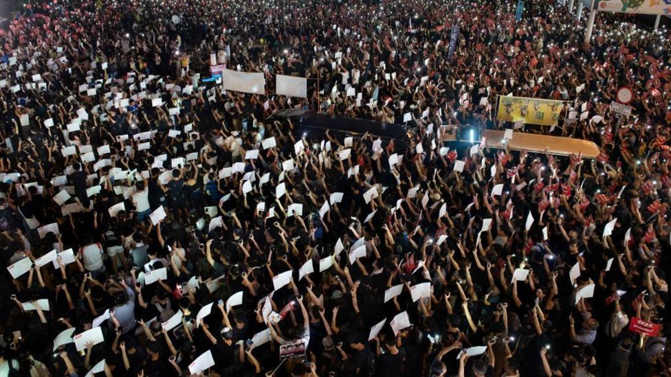 A crowd gathers in the night, raising banners