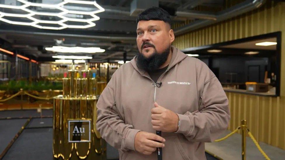 Charlie Sloth, wearing a beige tracksuit, stands in the foyer of the Au Vodka headquarters with a display of gold-coloured vodka bottles in the background.