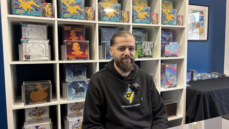 A man with dark hair tied back and a long, dark beard, dressed in a black hoodie smiles at the camera. He is sitting in front of shelving blocks, all of which contain a sealed casing of Pokémon cards or merchandise.