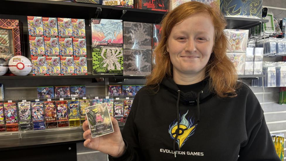 A young woman with pale skin and ginger hair, dressed in a black hoodie, smiles at the camera. She is holding up a Pokémon card with a small, pink creature on it and is standing in front of a shop's display of Pokémon cards.