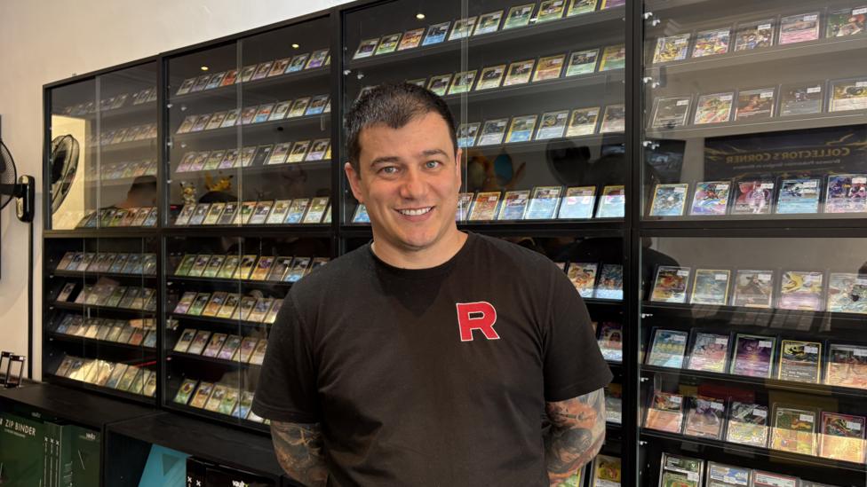 A man with dark hair and tattoos down both arms smiles at the camera. He is wearing a black T-shirt and is standing in front of shop cabinet of Pokémon cards.