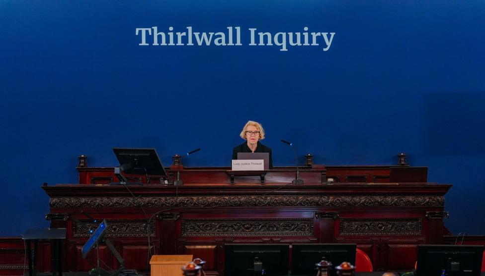 Image shows Lady Justice Thirlwall sitting on the raised bench at Liverpool Town Hall, with a large blue sign behind her that has 'The Thirlwall Inquiry' written in white letters.