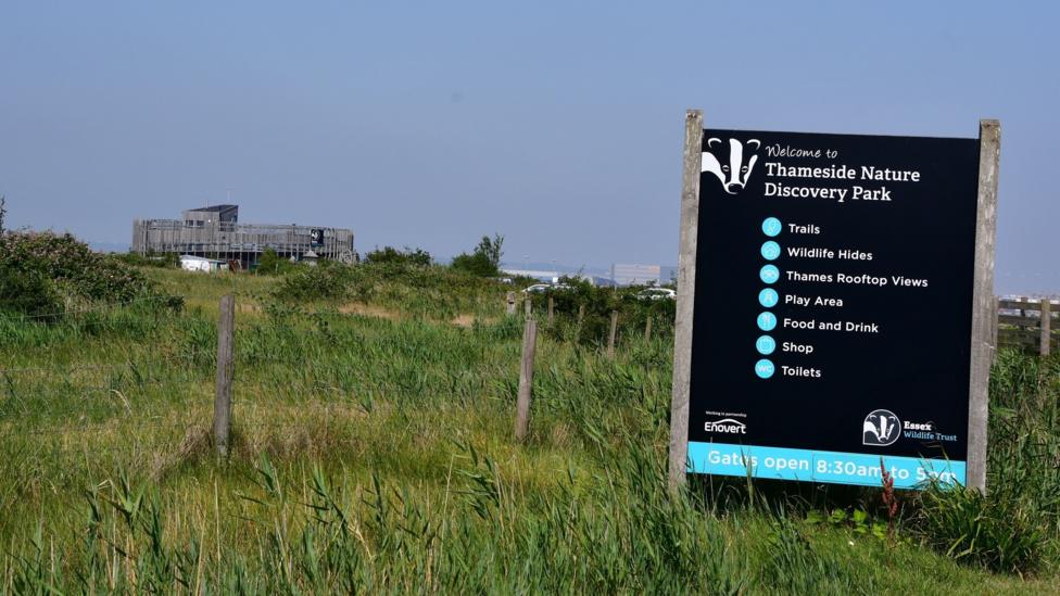 In the foreground is a black sign, with white-coloured font, which reads THAMESIDE NATURE DISCOVERY PARK. It sits on long grass. A blue hazy sky is above.