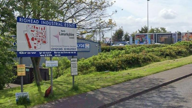 A sign for Righead Industrial Estate. It sits next to the pavement and there are units and billboards in the background.