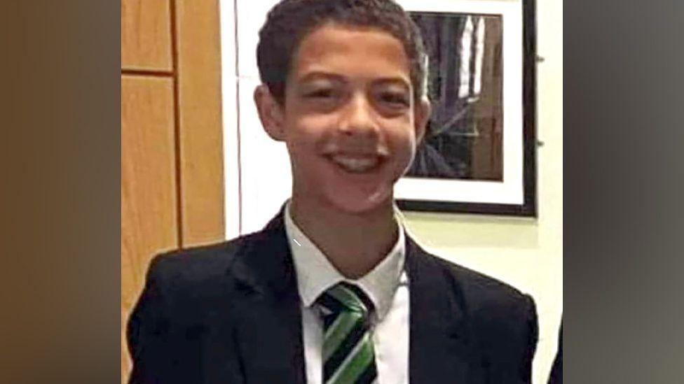 Noah Donohoe, a boy with dark hair, wearing a black blazer, with a white shirt and green, black and white tie.