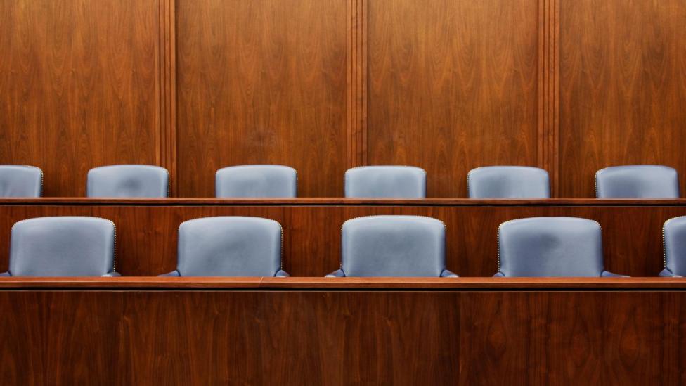 Empty jurors seats sit half hidden behind two long desks in a juror's box. The box is wooden and the seats appear leather.