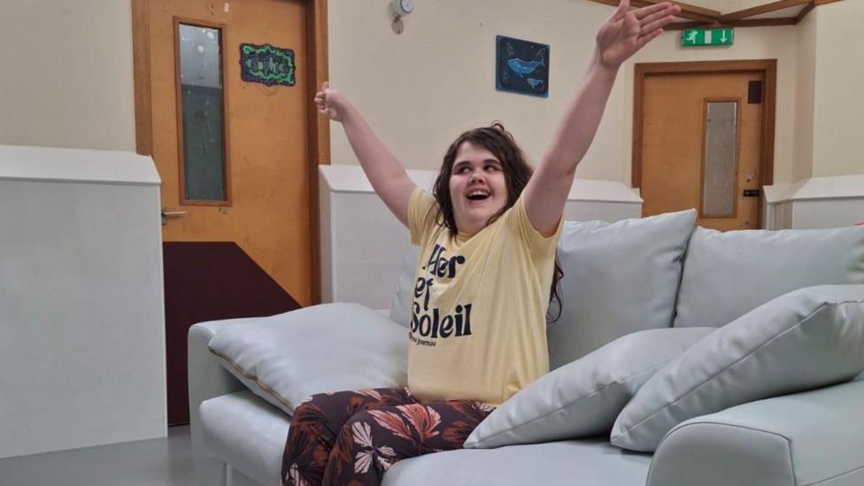 Sophie sitting on a couch wearing a pale yellow t-shirt and floral trousers. She has her arms outstretched and is smiling.