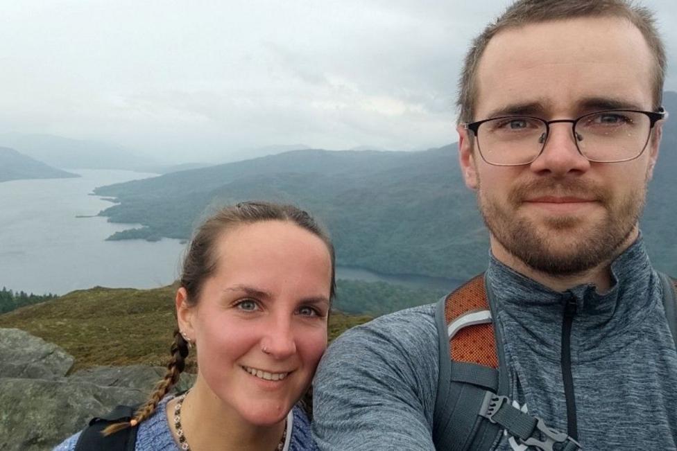 A woman and man pictured on top of a hill, taking a selfie. The woman has long brown braided hair, the man has short fair hair, a beard and glasses