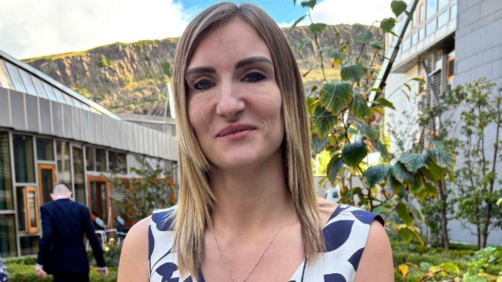 A woman with blonde hair in a close-up shot. She is wearing a patterned dress and standing in a garden with hills in the background