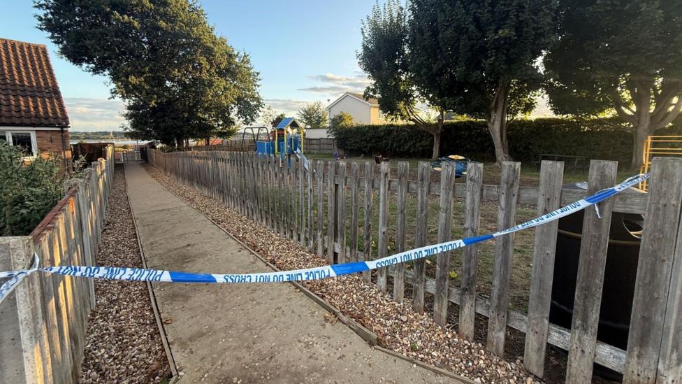 Blue and white police tape cordoning off a pathway next to a small park, which is lined with trees and a bush.