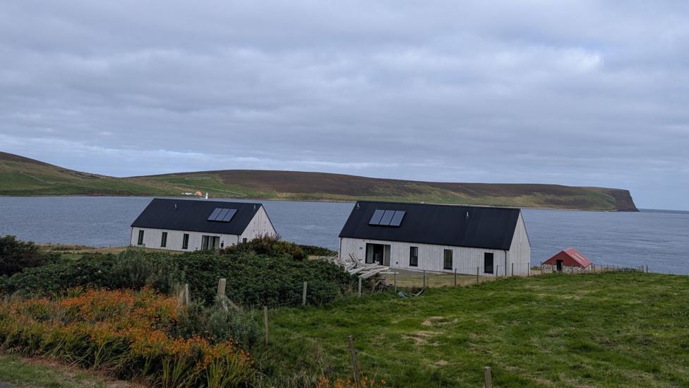 Two houses on the edge of an island. There is sea beyond them and a big cloudy sky. 