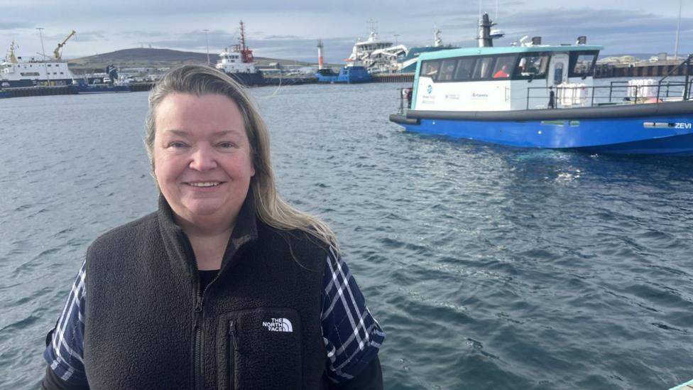 Woman in a black North Face jacket, standing at a harbour. There are a number of boats on the water behind her. 