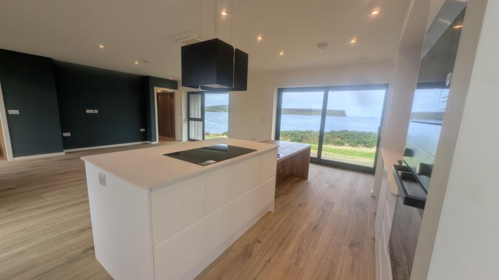 Interior of a new house. There is a kitchen island and a wooden floor. The view from the window is of another island and the sea. 