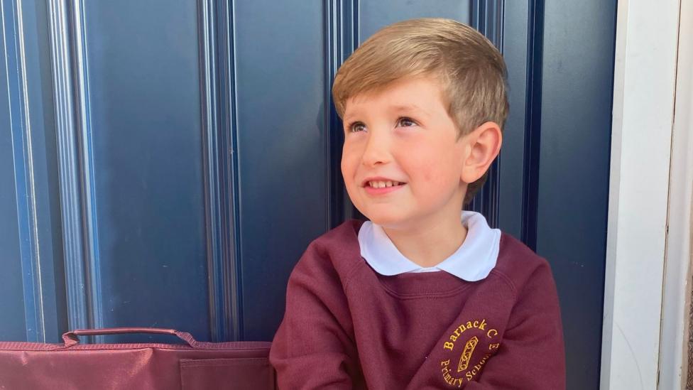 A boy in a maroon school jumper and white shirt looks to the left of the camera. It is a head and shoulders shot. He is in front of a dark blue door.