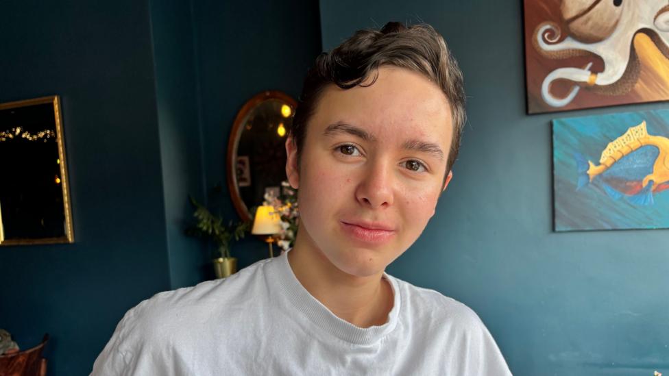 A head and shoulders shot of a boy with short brown hair looking at the camera. He has a white t-shirt on.