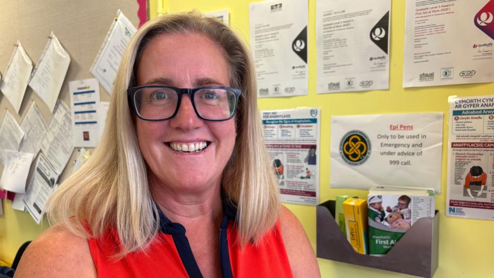A woman with blonde shoulder-length hair looking at the camera. She has glasses on and smiles. There is a wall with papers plastered on it - it looks like a school classroom or hallway.