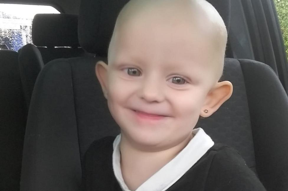 A little Kizzi-Rae sits inside a car and smiles towards the camera. Kizzi-Rae has no visible hair and is wearing a black short-sleeved top with a white collar and white sleeve trim. A small stud earring is visible in one ear. Dark car seats and windows can be seen in the background.