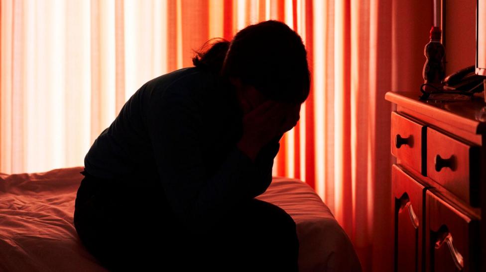 A woman in silhouette sits on top of a bed with her head in her hands