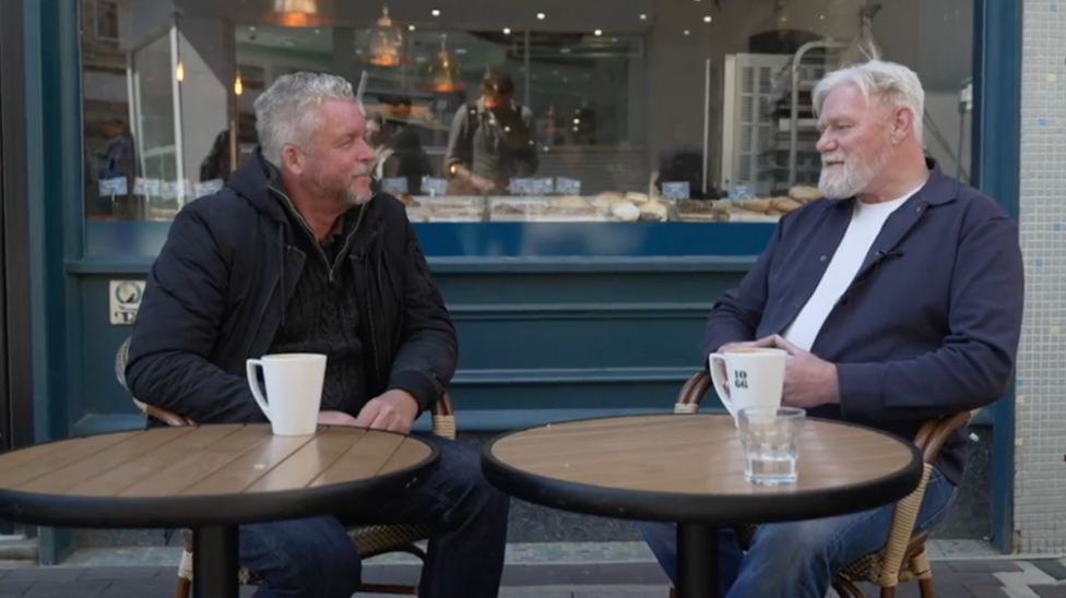 Two men sit at a coffee table. They are both wearing jackets and have grey hair. The man on the left has a goatee and the man on the right has a beard. 