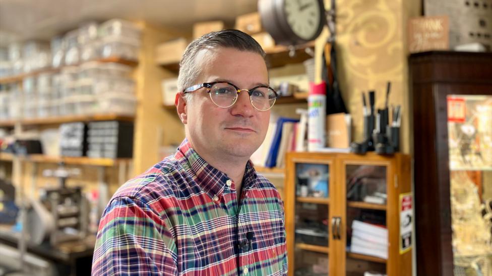 Man with glasses standing in a jewellery workshop. He is a wearing a green, red and purple plaid shirt.